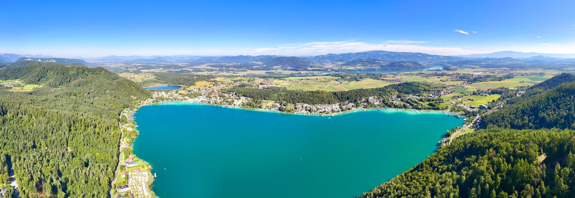Luftaufnahme des Klopeiner Sees: türkises Wasser, bewaldetes Ufer und Hügel unter blauem Himmel